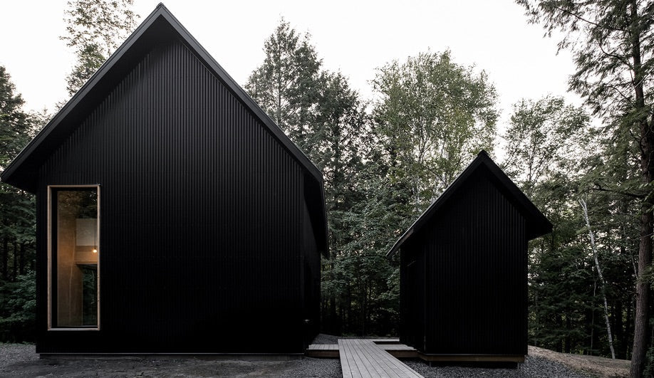 Clad in sleek black corrugated metal, this pair of cabins in the Quebec wilderness is linked by a cedar walkway